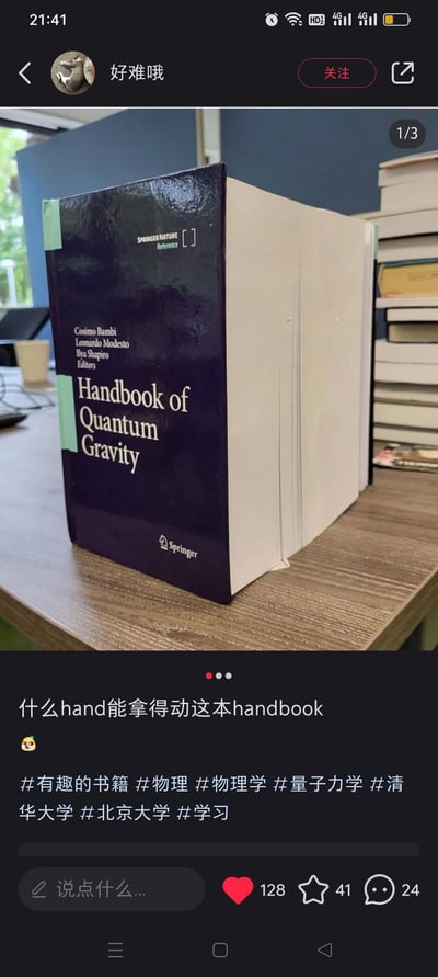 图片为社交媒体截图，展示了一本名为《Handbook of Quantum Gravity》（量子引力手册）的超厚学术书籍，放置在木质桌面上，旁边堆叠着多本其他书籍。书籍封面为深蓝色，标题文字清晰可见，整体厚度惊人。配文“什么hand能拿得动这本handbook”巧妙运用谐音梗，将“handbook”拆分为“hand”（手）和“book”（书），吐槽书籍过重难以手持，充满幽默调侃意味。适合学生或学术圈人士用于表达对厚重专业书籍的无奈与趣味分享。