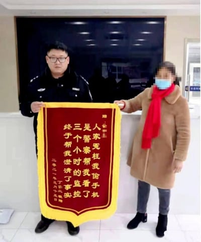 在派出所服务大厅内，一位身着警服的民警与一位戴口罩、穿驼色大衣配红围巾的女士共同举着一面锦旗。锦旗为红底黄字，文字口语化且真诚：“人家冤枉我偷手机，是警察帮我看了三个小时的监控，终于帮我澄清了事实”，落款为二零二一年三月十五日丁崔凤敬赠。这是女士为感谢民警帮她洗清被冤枉偷手机的嫌疑特意送来的锦旗，场景真实接地气。