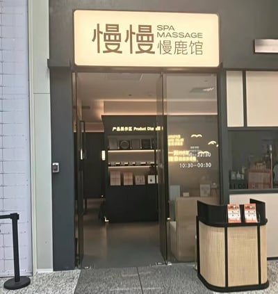 这是一张线下SPA按摩门店的门头实拍照片，店铺发光招牌上印有“慢慢”两个大字，右侧标注有“SPA MASSAGE 慢鹿馆”字样，组合起来店名为“慢慢慢鹿馆”，谐音“慢慢慢撸馆”契合按摩店的服务属性，带有趣味性。店铺入口为透明玻璃推拉门，玻璃上印有营业时间为10:30到次日00:30，向内可看到店铺内部设置有产品展示区，陈列着相关产品，门口右侧摆放了藤编材质的宣传展架，上面放置有宣传物料，整体装修风格简约舒适。