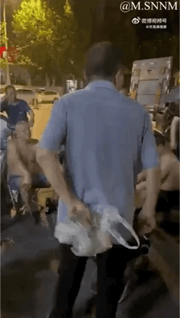 夜晚的街头，一名穿蓝色短袖衬衫的男子背对着镜头，双手拎着透明塑料袋，因视觉错位效果，看起来像是他的手被‘装’进了塑料袋内，画面充满趣味性。周围还有几位光着上身的市民坐在一旁，尽显生活化的市井氛围。