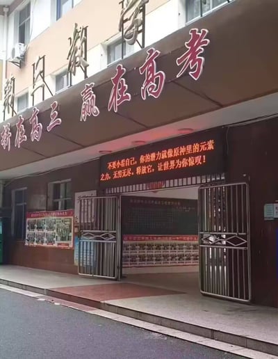 某高中高三年级教室入口处场景，建筑外墙上悬挂着红色大字标语“拼在高三 赢在高考”，下方电子屏则创新性地结合热门游戏《原神》元素，打出“不要小看自己，你的潜力就像原神里的元素之力，无穷无尽。释放它，让世界为你惊叹！”的励志文案。门口两侧为金属栅栏门，左侧墙面贴有学生活动照片海报，整体呈现出传统备考氛围与年轻流行文化的趣味融合。