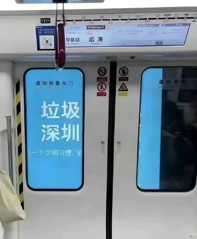 这是在深圳地铁11号线后海站车厢内拍摄的场景，车门处的电子提示屏被恶意篡改，原本的文明提示内容被替换为带有负面情绪的“垃圾深圳”字样，屏幕上方仍保留官方的“请勿扶靠车门”提示，下方还能看到原本文明提示语的残留“一个文明习惯，留”。车门区域贴有禁止吸烟、禁止饮食、小心夹手等安全标识，顶部的线路显示屏清晰显示当前为11号线后海站，属于停靠站状态。右侧车门玻璃内能看到模糊的乘客身影，整体场景反映了公共设施被恶意篡改的不文明行为。