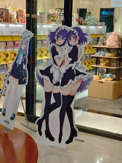这是在上海一家动漫主题店铺内的实拍场景，画面中有两款二次元角色等身立牌：左侧是手游《碧蓝档案》的高人气角色砂狼白子，身着校服，带有标志性兽耳与光环；右侧是标注有©Kyockcho,Karasumaya Presents的原创女仆双子，紫发异色瞳的两人亲密牵手依偎，姿态亲昵。背景可见店内的抓娃娃机、摆满毛绒周边与ACG商品的货架，整体充满浓厚的二次元氛围。
