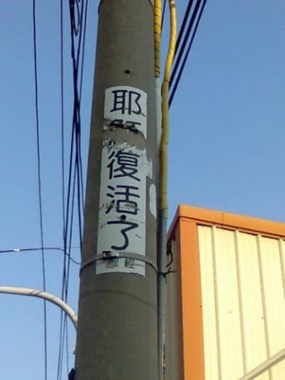 这是一张拍摄于街头的照片，内容是一根电线杆上贴着一张白底黑字的标语纸条，写着“耶稣复活了”。标语纸条有破损痕迹，但主要文字依然清晰。该画面通常用于凸显宗教标语与生活空间的强烈反差，有一种莫名幽默和无奈感，能引发网友吐槽或调侃。图片没有明显的双关或谐音梗。