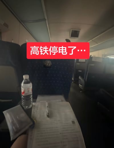 这张照片拍摄于高铁车厢内部，因高铁停电整体环境偏暗。前景是座位前方的小桌板，上面摆放着一瓶矿泉水、打印好的纸质材料、一支笔以及小包装袋，左下角露出了拍摄者的部分手部，后方可见高铁车厢的座椅和内部结构。画面上方叠加了红底白字的标注“高铁停电了…”，结合标题的调侃，暗指拍摄者原本可能要在行程中处理工作，遇上停电反而被迫获得休息，有种巧合的趣味感。