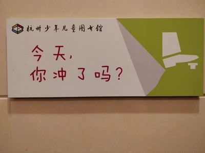 这是杭州少年儿童图书馆内的一块卫生间提示牌，采用浅白色与绿色拼接的版面设计，左上角印有杭州少年儿童图书馆的标识与名称，右侧配有冲水马桶的简笔画图案，版面中央用醒目的红色字体写着“今天，你冲了吗？”。提示牌用轻松俏皮的语气来引导使用者保持卫生间卫生，没有双关、谐音梗等内容，整体风格贴合儿童场所的活泼氛围，直白又亲切地传递了文明如厕的理念。