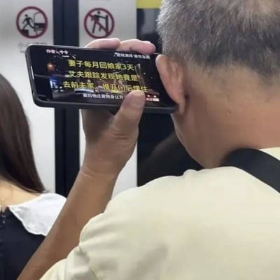 一位头发花白的老年男性在疑似地铁或车站的公共场所，正将智能手机贴在耳边并专注观看屏幕。手机屏幕上显示着黄色和白色的醒目文字，内容为具有戏剧性的社会新闻标题：“妻子每月回家3天 丈夫跟踪发现她竟是去前夫家，推开门后愣住 富哥你还要她让开”。背景中可见带有禁止进入图案的标识，周围还有其他模糊的行人。