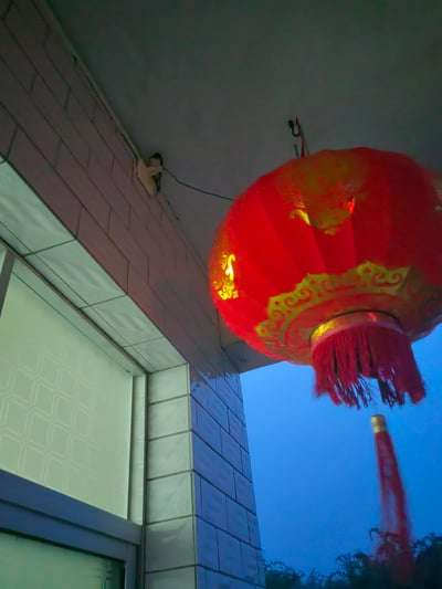 这是一张傍晚时分的实拍竖图，画面核心是悬挂于民宅屋檐下的红色中式灯笼，灯笼带有金色吉祥纹饰与福字类字样，内部亮着暖光，下方垂着红色流苏。背景是渐变为深蓝色的黄昏天空，旁边是带有瓷砖装饰的民宅墙面与窗户，整体氛围温馨祥和，充满中式传统节日的氛围感，图中无谐音梗、双关或暗示内容。