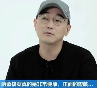这是一张来自游戏采访的视频截图，图中人物为《蔚蓝档案》的相关制作人，他戴着灰色鸭舌帽与细框眼镜，身穿黑色上衣，表情一本正经地看向画面一侧。图下方配有字幕“蔚蓝档案真的是非常健康、正面的游戏...”，由于《蔚蓝档案》游戏内存在不少偏向软色情的角色设计与剧情内容，这句官方说辞和实际游戏内容形成强烈反差，成为玩家用来调侃玩梗的素材。