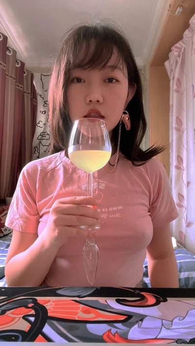 一位年轻女性在室内环境中自拍，她穿着粉色短袖T恤，左手手持一个装有黄色液体的高脚杯。女孩留着中长发，戴着一对带有链条装饰的耳环。背景中可以看到带有花纹的窗帘和房间内的部分家具，整体氛围显得轻松休闲。