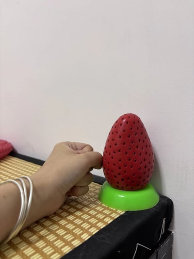 一只戴着银色手镯的手正在触摸一个草莓形状的红色物体，该物体表面有黑色圆点纹理，底部为绿色底座。物体放置在竹编纹理的垫子上，背景是白色墙壁，左侧隐约可见红色物体边缘。