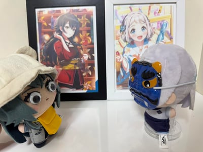 这是一张二次元偶像IP《偶像大师 闪耀色彩》的周边实拍展示图。前景是两款官方毛绒玩偶：左侧是戴着米色渔夫帽的浅仓透毛绒，替换了活动式大眼珠，造型呆萌；右侧是戴着蓝色鬼面具的久川飒毛绒，搭配透明支架摆放。背景是两幅装裱好的官方角色插画：左侧黑框画中是身着和风服饰的黛冬优子，手持苹果糖，背景暖色调风车装饰营造出温馨氛围；右侧白框画中是夏日造型的芹泽朝日，手持蓝色水炮，表情活泼开朗。整体画面呈现出温馨可爱的二次元收藏氛围，是爱好者的个人周边展示。