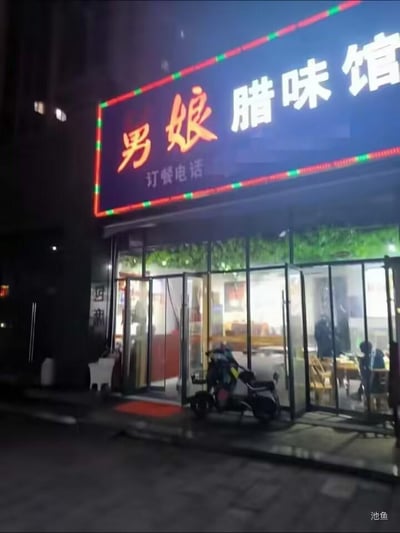 这是一张夜晚拍摄的街边餐馆外景照片，画面核心是名为「男娘腊味馆」的餐馆，招牌带有红绿相间的霓虹灯边框，下方标注“订餐电话”但号码被遮挡。餐馆的玻璃门处于敞开状态，门口停放着一辆电动车，地面铺有红色进门地垫；店内亮着暖光，能看到有顾客正在餐桌前用餐，整体呈现出夜间街边餐馆的日常营业氛围。招牌中的“男娘”字样较为特殊，容易引发趣味联想，成为画面的趣味亮点。