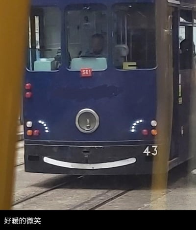 这是一张充满治愈感的视觉错位拍摄图，画面主体是一辆蓝色的有轨电车，电车底部的白色弧形条与黑色车身底部、中间的圆形部件，恰好组合成了一个微笑的拟人化表情，搭配下方的配文“好暖的微笑”，让原本冰冷的交通工具显得温馨又可爱，通过日常场景中的巧合，传递出轻松治愈的氛围。