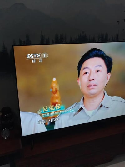 这是一张用户拍摄电视屏幕得到的翻拍截图，画面来自CCTV1综合频道的非遗相关节目。图中是官式琉璃烧制技艺协作手艺人巨能操，他正面对镜头做自我介绍，屏幕下方字幕显示“我是巨能操”，同时画面中也标注了他的身份信息。由于名字的谐音容易引发趣味联想，成为这张图的核心亮点，节目本意是宣传传统非遗技艺与手工艺人。