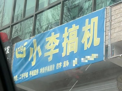 图片展示了一个悬挂在建筑物外的蓝色横幅招牌，上面用黄色大字写着“小李搞机”，下方小字标注主营项目：“二手机 手机配件 冲洗手机照片 打字 复印”。背景可见玻璃幕墙建筑和光秃秃的树枝，推测为冬季或初春时节拍摄的街头店铺实景。