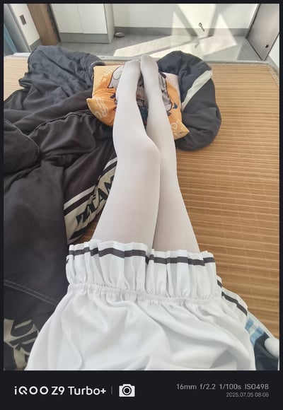 这是一张第一视角的个人自拍图，展示了穿着白色连裤袜与带黑边装饰白色睡裙的腿部，腿部搭在印有动漫图案的抱枕上，背景是铺有竹席的床铺，旁边还有深色的被子，整体呈现出卧室里的慵懒日常氛围，穿搭风格偏向二次元软妹风。
无双关、谐音梗或暗示内容。
