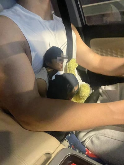 A person driving a car while wearing a white tank top that features a printed image of another person with a blurred face. The driver is holding a yellow stuffed toy in their left arm while operating the vehicle with their right hand. The image appears to be taken from the passenger seat perspective, showing the driver's upper body and the car interior.