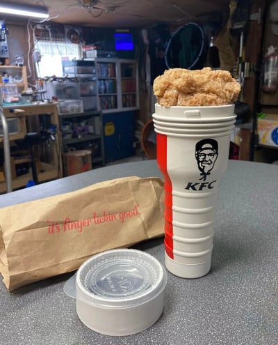 图片展示了一个创意场景：一个红白相间的KFC纸杯被装满了金黄酥脆的炸鸡块，杯身上印有经典的山德士上校头像标志。杯子旁边放着一个印有"it's finger lickin good"标语的棕色纸袋和一个带盖的透明塑料小容器。背景是一个略显杂乱的 workshop 环境，有工具架、储物柜和各种设备，营造出一种随意而有趣的氛围。这种将炸鸡装入饮料杯的非常规吃法形成了视觉上的反差萌，突出了创意美食的主题。