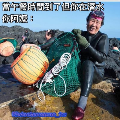 图片中一位穿着黑色潜水服的阿姨，戴着粉色边框潜水镜，正坐在海边的岩石上，双手紧握一个绿色的渔网，身旁放着一个橙黄色的浮标。她面带灿烂的笑容，背景中还有其他穿着潜水服的人在岩石间活动。图片上方配有文字“當午餐時間到了但你在潛水 你阿姨：”，幽默地将午餐时间与潜水场景结合，暗示阿姨潜水归来可能带回了海鲜午餐，整体氛围轻松有趣。