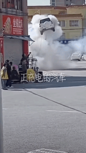这是一段动态GIF图，呈现了街头的惊险突发场景：一辆正在充电的汽车意外发生爆炸，强大的冲击力将汽车直接炸至腾空飞起，现场冒出大量白色浓烟。街边店铺门口的路人被这突如其来的一幕惊到，纷纷驻足看向爆炸方向。图中配文“正充电的汽车”点明了事件的起因，画面极具冲击力，直观展现了电动车充电意外的危险性，场面既离谱又让人感到后怕。