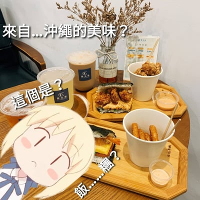 圖中展示了一張木桌上的美食擺盤，包含兩個竹製托盤，分別盛放著不同食物：左側托盤有一杯黃色飲料和一杯棕色奶茶（均貼有「暖九屋」標籤），右側托盤有炸雞塊、帶有炸物內餡的飯糰，以及另一盤炸條狀食物，旁邊配有小杯蘸醬。背景中有一個插著乾花的玻璃瓶和一份菜單。左下角貼有一個卡通少女形象，她金髮、閉眼、臉頰泛紅，表情疑惑，周圍配有文字「來自...沖繩的美味？」「這個是？」「飯...糰？」，表達對眼前食物的好奇與不解。