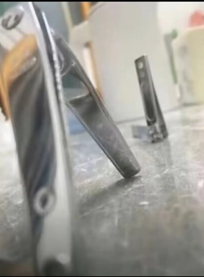 图片展示了一个 workshop 环境中的金属工具特写。前景中有一个较大的金属部件，具有三角形的镂空设计和螺栓孔，表面呈现出金属特有的光泽和纹理。背景中可以看到另一个较小的金属支架，同样具有类似的工业设计风格。整个场景似乎是在一个工作台上拍摄的，表面有使用痕迹和划痕，暗示这是一个活跃的金属加工环境。