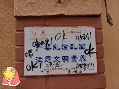 这是一张拍摄于上海虹口区的线下宣传标语牌照片，标语牌原本是官方用于引导市民文明行为的宣传物料，核心内容为“严禁乱涂乱画 注意文明素养”，同时标注了“全力创建全国文明城区”“社会主义核心价值观”相关内容，落款为四川北路街道党工委、办事处，还有“HongKou 虹口”“川北景象”的标识。但该标语牌被人用黑色马克笔大面积乱涂，涂鸦内容包括“okay!OK”“收到!”“喂”“ok!下次一定”“收到!!!”等字样，完全违背了标语牌本身的要求，形成了强烈的讽刺反差。照片左下角还添加了一个捂嘴坏笑的卡通表情，进一步强化了这种讽刺戏谑的氛围，凸显了行为与标语要求的矛盾，讽刺了明知故犯的不文明行为。
