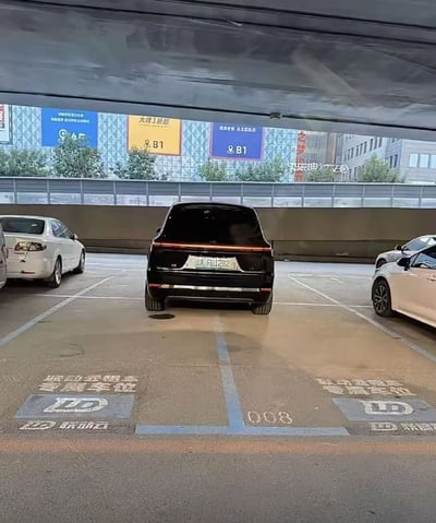 停车场场景图，画面中央一辆黑色汽车正停放在标有“联动云租车 专属车位”及编号“008”的停车位上。车位地面有白色文字标识和“LD” logo。左右两侧分别停放着白色轿车，背景可见建筑物外墙及多块广告牌，其中黄色广告牌显示“大连上新品”，蓝色和紫色广告牌标注“B1”区域。整体场景为日间户外停车场，光线自然，构图清晰。