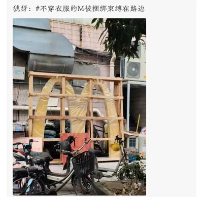 图片展示了一个被木板框架固定的麦当劳黄色M标志，外面包裹着塑料布，周围有自行车和老旧建筑物背景。上方配有文字'猇犽：#不穿衣服的M被捆绑束缚在路边'，通过双关语制造笑点：'不穿衣服的M'指被塑料布包裹的麦当劳标志，'捆绑束缚'则形容其被木板框架固定的状态。