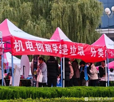 这张图片拍摄的是大学校园户外社团招新活动现场，前景是修剪整齐的绿色绿篱，背景是枝叶茂密的垂柳，场地上搭建了多个粉白相间的招新帐篷。其中一个帐篷悬挂的红色横幅因文字排版拼接失误，原本应为“机电-创新学会 垃圾分类”的横幅内容，被错误排版断句成“机电-创新学会是垃圾 分类”，产生了极具反差感的搞笑歧义，多名学生在该招新摊位周边活动，图片右下角标注了来源水印“@山东理工大学吧”。