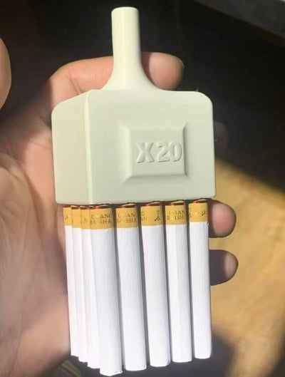 一只手握着一个浅绿色的矩形装置，装置顶部有一个圆柱形凸起，正面中央刻有“X20”字样。装置下方整齐排列着7支香烟，每支香烟的过滤嘴为黄色，上面印有“CANG BAI SHA”的字样及图案。背景较暗，手部和装置成为视觉焦点，整体呈现出一种新奇的物品组合场景。