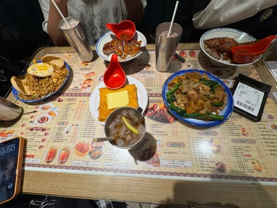 图片展示了尖沙咀红茶餐厅的聚餐场景，餐桌上摆放着多道美食，包括两碗烧鸭饭（配红色小勺子）、一盘煎蛋炒饭、一盘炒牛河（含青菜和肉片）、一份表面放有黄油的西多士，以及多杯插有吸管的不锈钢杯装饮品和一杯带柠檬片的冰饮。桌面铺有印有菜品图片和中文文字的菜单式桌布，桌边可见用餐者的手臂和部分衣物，整体呈现出轻松日常的聚餐氛围。