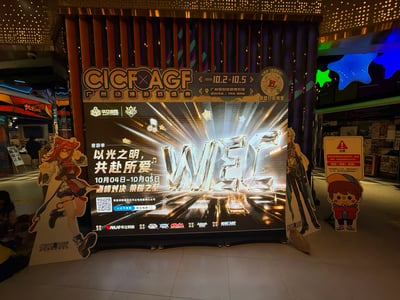 照片拍摄于光线偏暗的商场内部，主体是一处CICF×AGF广州动漫游戏盛典的线下宣传展位。展位中央为大型黑色发光展板：顶部金色标题栏写有“CICF×AGF 广州动漫游戏盛典”，标注展会时间为2025年10月2日至10月5日，地点为广州保利世贸博览馆，邻近地铁8号线/11号线琶洲站，并提示微信扫码购票，标题栏右侧还有圆形倒计时风格图标。展板主视觉属于华立电竞WEC音游季，中央是水晶切割质感的巨大立体“WEC”字母，配以金色放射光线、音符和星星装饰，地面延伸出音游轨道灯效并出现“PERFECT”判定字样；左侧文案为“音游季 以光之明，共赴所爱 10月04日-10月05日 巅峰对决 荣耀之至”，下方设有二维码、“公众号搜索 华立电竞”搜索框、“赛事详情请关注华立电竞赛事公众号”提示，底部标注“赛事主办 WAHLAP华立科技”及多个赛事合作品牌logo。展板左侧立有红发、红黑配色服饰的二次元少女等身人形立牌，呈动感跳跃姿势，底座印有CICF×AGF字样；右侧立有浅色头发、穿长款外套的高个男性角色立牌，其旁是戴黄蓝拼色棒球帽、穿红外套、双臂交叉的Q版小男孩提示牌，更右侧立有一块带红色三角警示标识的“注意”告示牌。背景可见商场的古铜色与深蓝色竖条装饰墙、镂空点阵吊顶、蓝绿星形灯饰，左侧远处有“机迷会”店铺和展架，还有一名黑衣行人。整体为真实线下实拍，具有浓厚的电玩与漫展宣传氛围。