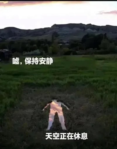 这是一张充满浪漫想象力的创意梗图，背景为黄昏时分的郊野景观：上方天空残留着粉橘色的落日霞光，远处是层叠的暗色调山脉，中部是连片的深绿色草地，草地下方位置有一个呈大字型展开的人形凹陷，凹陷表面反射着天空的霞光，看起来仿佛是天空化成人形躺进草地里休息。图中搭配两行白色粗体文字，上方配文为‘嘘，保持安静’，下方配文为‘天空正在休息’，整体氛围松弛治愈，充满拟人化的趣味脑洞。