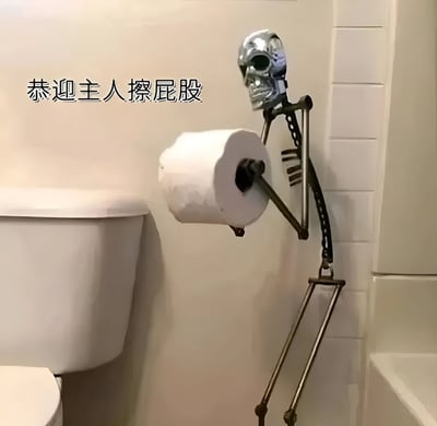 浴室场景中，一个造型独特的骷髅形状卫生纸架安装在马桶旁边的墙上。骷髅头部朝向马桶方向，一只金属骨架手臂托着一卷卫生纸，姿态仿佛在恭敬地等待使用者。图片左上角配有文字“恭迎主人擦屁股”，与骷髅卫生纸架的造型形成幽默对比，营造出一种滑稽搞笑的氛围。
