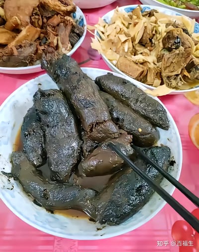 图片展示了一桌丰盛的家常菜，中心是一个白色瓷碗，里面盛放着深色、油亮的卤味，可能是卤猪手或类似食材，表面覆盖着浓稠的棕色酱汁，筷子正从碗中夹起一块。碗的周围摆放着其他菜肴：左侧是一盘棕色的炖肉，右侧是一碗混合了笋干和腐竹的炖菜，背景中还能看到部分绿色蔬菜。整张桌子铺着粉色带有花纹的桌布，整体氛围像是家庭聚餐或节日宴席，食物看起来热气腾腾，色泽诱人。
