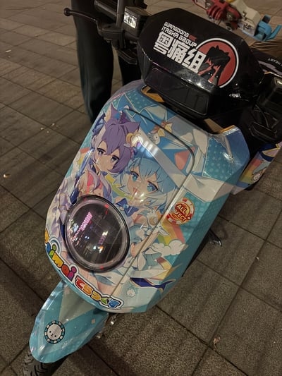 这是一辆经过精心改装的动漫主题电动车（痛车），车身以蓝色为基调，覆盖着色彩鲜艳的动漫少女角色图案。车头部分有一个醒目的圆形大灯，灯上方印有"雪痛组"和"SHOGUN TASHA GROUP"字样的黑色标志，标志中还包含一个动漫人物剪影。车身侧面可以看到"imai"字样和多个动漫元素贴纸，整体设计充满了二次元风格。车辆停放在铺有灰色地砖的地面上，背景中能看到部分路人的腿部。