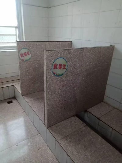 这是在一间贴满白色瓷砖的房间内，将原本类似卫生间蹲位的平台区域，改造成了带有花岗岩质感隔板的独立就餐位，隔板上贴着带有装饰图案的“就餐位”标识。这种将如厕功能区域改造为就餐区的设计充满强烈反差，显得十分荒诞猎奇。