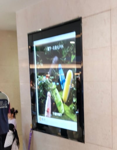 这是一张在商场公共区域拍摄的竖版电子广告屏照片，屏幕展示着蕉下品牌的轻量化户外产品——多款色彩鲜亮（粉、绿、黄、蓝、灰）的胶囊式折叠伞，背景搭配绿植场景，上方标注文字“蕉下·轻量化户外”，下方则标明店铺位置“6楼F2号蕉下”。屏幕左侧可见几位路人的身影，周围墙面为浅色石材，整体环境是装修精致的购物中心通道。