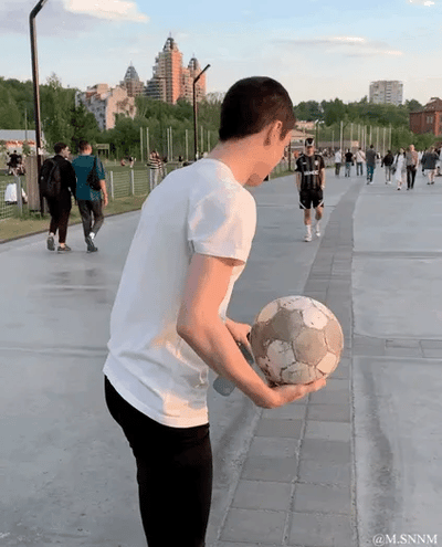 这是一个动态GIF图，在户外的城市休闲步道上，一名穿白色短袖、黑色长裤的男子双手捧着一个旧足球，通过巧妙的肢体摆放和拍摄角度，形成了有趣的视觉错位效果：看起来他的手臂仿佛和足球融为一体，像是手部是足球的一部分，制造出荒诞又搞笑的错觉。背景中还有不少行人，远处可见高楼和绿植，整体场景是日常的户外环境。