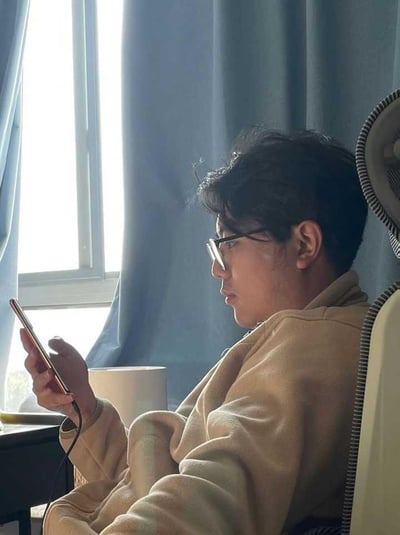 图片中，一个男子穿着宽松睡衣或家居服，懒洋洋地半靠在椅子或床上刷着手机，身旁有一个白色杯子和窗帘被阳光照亮，整体氛围非常随意舒适，给人一种宅家、休闲的感觉。画面侧面突出安逸和慢生活状态，可能暗示当代年轻人居家时间变多、喜欢赖床玩手机等现象。没有明显的双关、谐音梗或暗示。