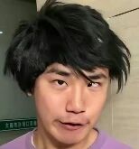 图片为一名男子的面部特写，他有着凌乱的黑色短发，身穿紫色上衣，表情看起来有些惊讶或困惑。背景是室内浅色墙面，左侧隐约可见绿色文字标识（内容不清晰）。整体画面为静态单图，无明显文字内容或水印。
