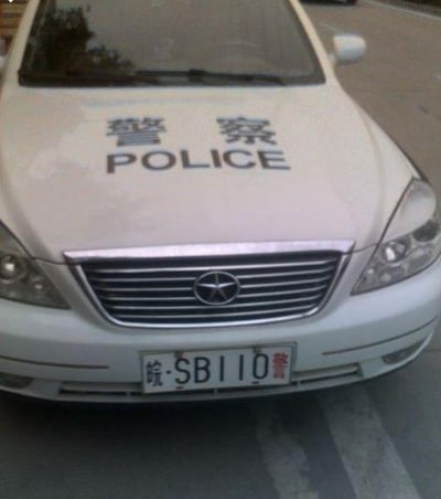 图片展示了一辆白色警车的正面特写，车头引擎盖上印有“警察”及“POLICE”字样，前脸格栅中央有汽车品牌标识，车牌号为“皖·SB110警”。车辆停放在道路上，背景为模糊的路面。车牌中的“SB”组合在网络语境下常被视为带有幽默调侃意味的谐音梗（谐音“傻X”），使得该图片具有一定的搞笑效果。