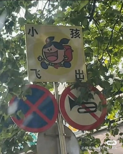 这是一张户外拍摄的道路标识组合图，画面被茂密的树叶部分遮挡。最上方是一块自制的提示牌，牌面中心是一个背着红色书包、咧嘴大笑的卡通小孩，做出奔跑跳跃的活泼动作，提示牌四个角分别标注“小”“孩”“飞”“出”四字，组合成“小孩飞出”的提示语，用以生动地提醒过往车辆，附近区域可能有儿童突然跑出，需注意避让。提示牌下方的金属杆上固定着两个正规交通标志，左侧是蓝底红叉的禁止车辆临时或长时停放标志，右侧是红圈白底、带有被划掉的喇叭图案的禁止鸣笛标志，两个标志均被绿植部分遮挡。