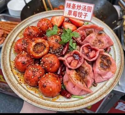 整张图展示的是一道猎奇创意混搭小吃，一个粗陶浅盘被人托着，盘内左侧是经红油炒制的汤圆，外皮呈红亮的油润色泽，表面撒有白芝麻、花椒、干辣椒段，部分汤圆被切开露出内馅，是辣条炒汤圆；盘内右侧是粉红色面皮的饺子，部分饺子被切开可以看到内部的草莓馅料，旁边还搭配了新鲜草莓切片和香菜叶作为点缀，盘上方立有白底红字的小标牌，标注菜品名称为“辣条炒汤圆 草莓饺子”，背景隐约可见小吃摊的其他食材和炊具，属于颠覆传统口味的黑暗料理类街头小吃。
