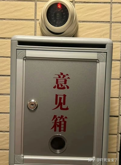 这张图片展示了一个安装在仿砖纹墙面上的银灰色意见箱，箱体正面印有醒目的红色“意见箱”字样。在意见箱的正上方，安装着一台处于工作状态的半球形监控摄像头，摄像头的红色指示灯亮起，明确显示正在拍摄。原本设计用于匿名收集公众反馈的意见箱，被监控设备直接对准，形成了极具讽刺性的场景——这种设置会让想要投递意见的人顾虑自身行为被记录，彻底消解了意见箱匿名收集反馈的核心意义，是对形式主义管理方式的生动调侃。