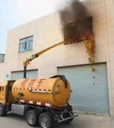 图片中展示了一辆黄色的吸粪车停在一栋米黄色建筑物前，车辆的长臂吸管伸入建筑物上层的一扇窗户内，正从窗中吸出大量深色浓稠的污物，部分棕褐色残留物顺着墙面滴落。建筑物底层有一扇关闭的卷帘门，吸粪车车身侧面可见红色文字。整个场景呈现出一种非常规且令人惊讶的作业画面，未见人员出现。