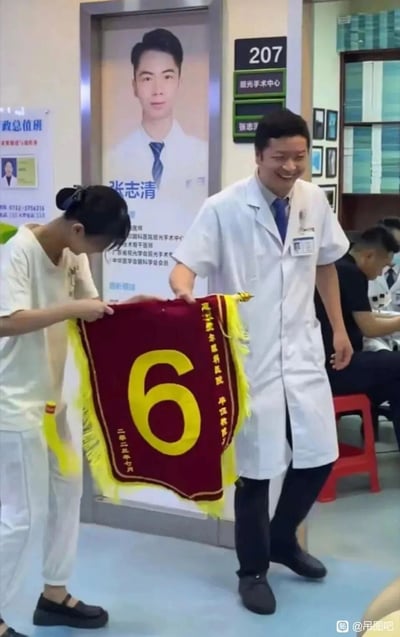 这张图片拍摄于湛江爱尔眼科医院的207屈光手术中心，画面里一位年轻女患者和一位穿着白大褂的男医生共同举着一面红底黄字的锦旗，锦旗上印有大大的数字“6”，竖排文字为“湛江爱尔眼科医院 单位敬赠”，落款是二零二三年七月。背景墙上张贴着医生张志清的个人介绍海报，而眼前的这位医生和海报上的张志清相貌明显不符，医生脸上露出尴尬又无奈的笑容，患者则低头显得十分窘迫，显然是患者认错了主治医生，送错了锦旗，现场氛围充满社死感。
