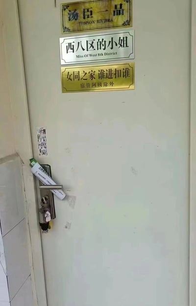 这是一张宿舍门的实拍照片，门上垂直排列着三张风格各异的门贴。最上方是金色边框的“汤臣一品 TOMSON RIVIERA”（知名 luxury 楼盘名），中间为白色边框的“西八区的小姐 Miss Of West 8th District”（疑似宿舍成员的趣味自称），最下方是金色边框的“女同之家 谁进扣谁 宿管阿姨除外”（直白表明宿舍属性并附带俏皮“警告”，特别豁免宿管阿姨）。门把手左侧贴有小幅贴纸，门把手上挂着钥匙串，整体呈现出大学生宿舍特有的创意与幽默感。